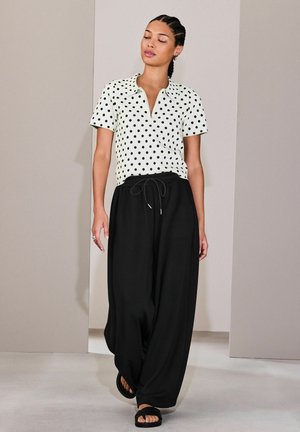 Woman with braided hair wears a white short-sleeve polka dot shirt, loose black pants, and black sandals, standing against a neutral background.