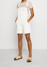 Bílé denimové overaly s ohrnutými šortkami, s nastavitelnými ramínky a přední kapsou. V kombinaci s pruhovaným tričkem a šlapkami.