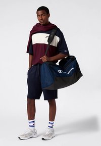 Granatowa torba typu duffel z czarnymi akcentami, z logo Champion. Model ma na sobie koszulkę w kolorze bordo i białe panele oraz granatowe szorty.