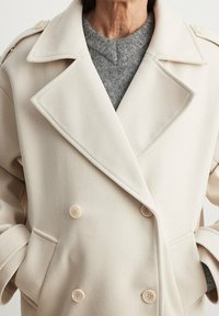 Personne portant un manteau croisé couleur crème sur un pull gris en maille, les mains dans les poches du manteau, visible du cou jusqu’aux hanches.