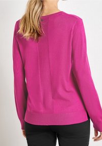 Kobieta nosząca magentowy sweter z długim rękawem i małym guzikiem z tyłu u góry, w połączeniu z czarnymi spodniami.