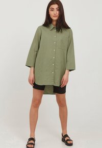 Camicia leggera verde oliva con colletto, maniche a tre quarti e taschino sul petto, abbinata a pantaloni corti neri e sandali.