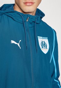 Teal takki hupulla, jossa on valkoinen Puma-logo ja Marseillen joukkueen vaakuna. Valmistettu sileästä, kevyestä materiaalista.