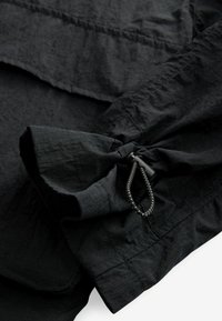 Schwarzes Kleidungsstück mit strukturiertem Stoff, das einen gerafften Ärmel mit Schleifendetail und verstellbarem Kordelzug für die Passform aufweist.