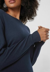 Marineblaues Sweatshirt mit weicher Textur, weiten Ärmeln und runder Ausschnitt. Nahaufnahme von Händen, die den Stoff lässig greifen.