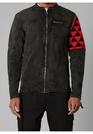 Uomo che indossa una giacca nera con zip con una toppa triangolare rossa e nera sul braccio destro superiore, abbinata a pantaloni neri.