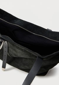 Μαύρη τσάντα χειρός από σουέτ με ασημένιο φερμουάρ, μαύρα δερμάτινα λουριά και ανοιχτό κύριο διαμέρισμα που αποκαλύπτει το σκοτεινό εσωτερικό ύφασμα.