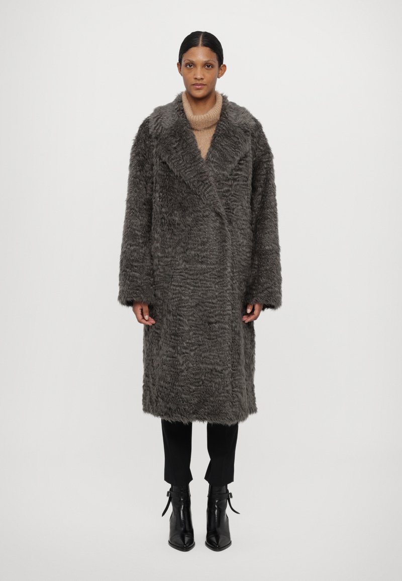 Pilkos dirbtinės kailio striukės su tekstūruota paviršiumi, plačiomis apykaklėmis ir ilgomis rankovėmis, dėvimos ant kreminio megztinio ir juodų kelnių.