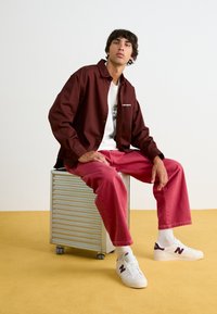 Burgundská košile na knoflíky přes bílý tričko s potiskem, v kombinaci s růžovými kalhotami a bílými teniskami s fialovými akcenty, sedící na stříbrném vozíku.