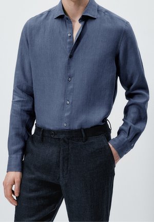 Homme portant une chemise bleue à manches longues boutonnée, rentrée dans un pantalon gris foncé, avec une main dans la poche et l'autre relâchée.