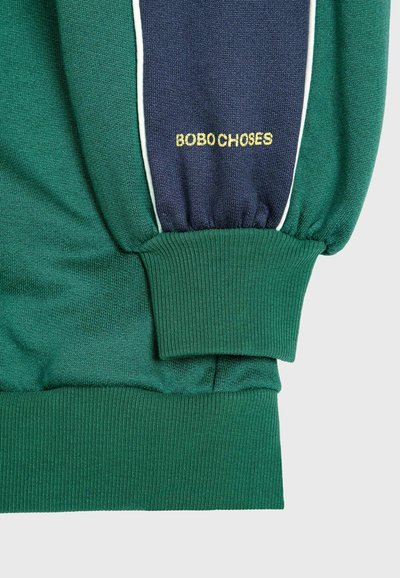 Zeleni dukserica s mornarskim bočnim panelima, elastičnim rukavima i bijelim obrubom. Ima zlatni vez s natpisom "BOBO CHOSES" na ruci.