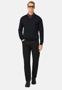 Pull noir à col en V porté sur une chemise marron à col, associé à un pantalon noir et des baskets noires. Le mannequin se tient avec les mains dans les poches.