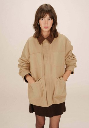 Veste oversize beige avec un col en velours côtelé marron, deux poches avant et un tissu texturé. Portée sur une robe marron.