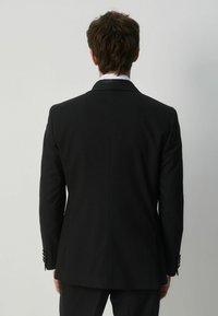 Mann mit braunen Haaren, der einen schwarzen Anzugjacke und ein weißes Hemd trägt, von hinten vor einem schlichten hellgrauen Hintergrund.
