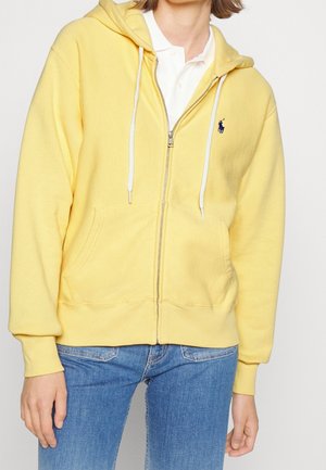 Person wearing a yellow zip-up hoodie with white drawstrings over a white collared shirt and blue jeans, standing against a plain background.