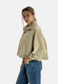 Beżowa krótka kurtka z szpiczastym kołnierzem, zapinana na guziki z regulowanymi paskami przy mankietach. Noszona z niebieskimi dżinsami, charakteryzuje się gładką fakturą.