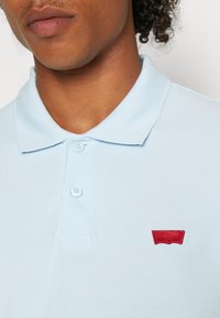 Ljusblå polo t-shirt med strukturerad tyg, med en treknappslåsning och en liten röd broderad logotyp på vänster bröst.