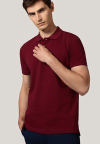 Burgundowa koszulka polo z krótkim rękawem, płaskim kołnierzykiem i zapięciem na trzy guziki. Miękka, gładka tkanina, dopasowany krój i szczegóły po bokach.