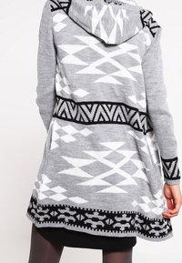 Grå stickad cardigan med huva, prydd med vita geometriska mönster och svarta accenter längs nederkanten och ärmarna. Avslappnad, knälång design.