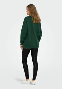 Sudadera verde de gran tamaño con cuello redondo, mangas largas y puños acanalados, combinada con leggins negros y zapatillas blancas.