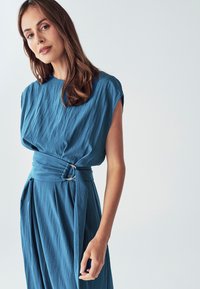 Zils, drapēts kleita ar īsām piedurknēm, viduklī nospiesta ar sudraba sprādzi, teksturētu audumu un plūstošu siluetu.