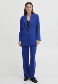 Modrý blazer s jedním knoflíkem, klopovým límcem a dvěma předními kapsami, sladěný s širokými kalhotami. Texturovaná látka, elegantní design.