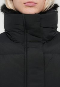 Crna podstavljena jakna s visokim ovratnikom obrubljenim umjetnim krznom, prešivanom teksturom i modernim dizajnom.