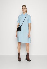 Vestido camiseiro azul claro, com mangas curtas e gola, combinado com botas de tornozelo castanhas e uma pequena bolsa tiracolo preta.