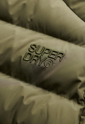 Olivgrüner Pufferjackenstoff mit genähter Struktur. Verfügt über das geprägte „SUPERDRY“-Logo in passendem grünen Faden. Glatte Oberfläche, leichter Glanz.
