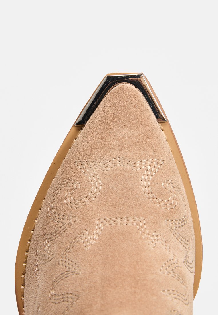 Botín de ante beige con punta afilada, con patrón western en relieve y un detalle metálico negro en la punta. Suela de cuero suave.