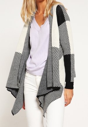 Kvinde iført en sort-hvid ternet cardigan med åben front over en lyse lilla V-hals sweater og hvide bukser.