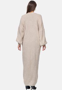 Long, beige knitted dress with oversized sleeves, ribbed texture, and a relaxed fit. Features a high neckline and falls to the ankles.