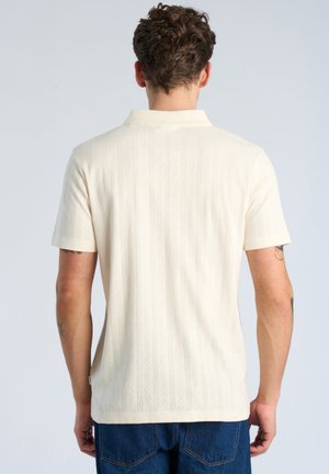 Mężczyzna z kręconymi włosami, ubrany w kremową, teksturowaną koszulkę polo z krótkim rękawem i niebieskie dżinsy, pokazany od tyłu.