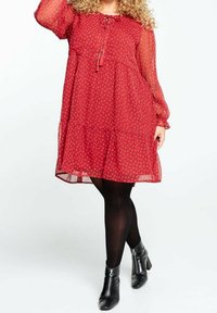 Robe rouge transparente avec des manches longues, un détail à nouer au cou, un ourlet à volants et de petits motifs floraux blancs. Portez-la avec des collants noirs et des bottines à zip.
