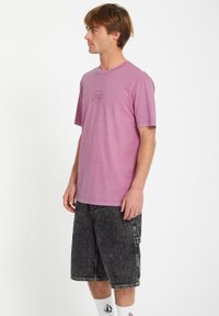 Lyslilla kortærmet t-shirt med rund halsudskæring og et diskret logo centreret på brystet, parret med mørkegrå denim shorts.