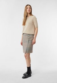 Beige gebreid T-shirt met korte mouwen, gecombineerd met een geruite kokerrok met een zijsplit en knoopaccenten. Model draagt zwarte schoenen en sokken.