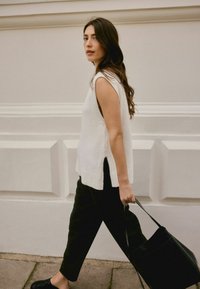 Biela pletená top bez rukávov so bočnými rozparkom, v kombinácii s čiernymi nohavicami. Modelka drží čiernu tašku a má dlhé, vlnité vlasy.