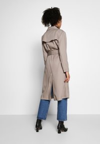 Trench coat beige con vita con cintura e ventaglio posteriore, abbinato a jeans blu e stivali neri con tacco. Tessuto liscio e design strutturato.