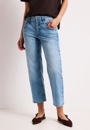 Lyseblå cropped jeans båret med sort skjorte og brune flade sko, stående med den ene hånd i lommen mod en ensfarvet baggrund.