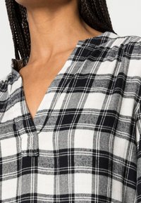 Chemise à carreaux noir et blanc présentant un col en V, des manches courtes et une texture de tissu douce. Le motif comprend différentes formes carrées.