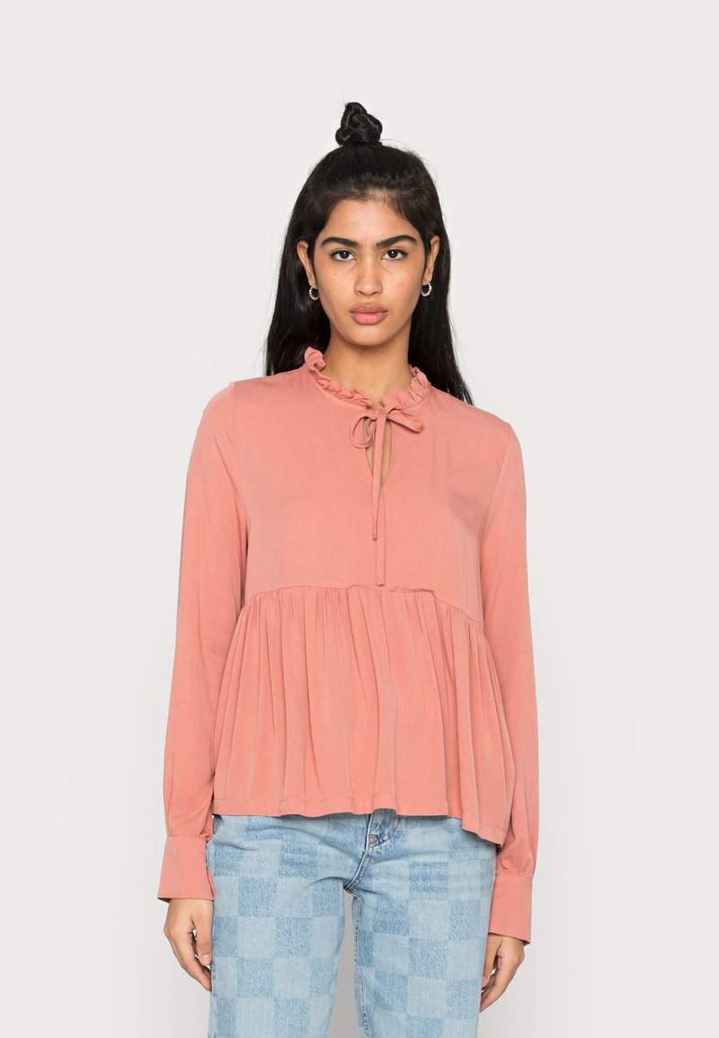 Blusa de manga longa, rosa claro, com colarinho com babados e detalhe de laço, apresentando tecido franzido abaixo do busto e uma textura suave e fluida.