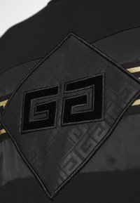 Schwarzes Kleidungsstück mit einem strukturierten, rhombusförmigen Patch, das ein geprägtes Logo und dezente geometrische Muster aufweist. Goldene Akzente verlaufen horizontal.