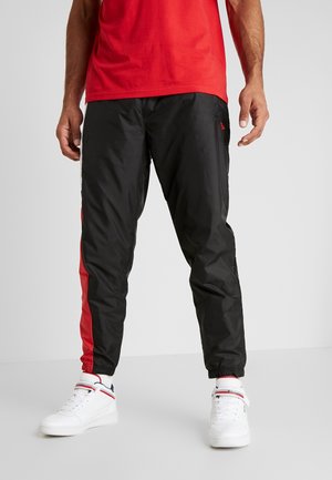 Man wearing black and red athletic pants with elastic cuffs and white sneakers, standing against a plain light background.