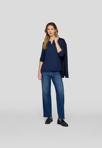 Dunkelblaue Bluse mit langen Ärmeln, rundem Ausschnitt und vorderen Taschen, kombiniert mit weit geschnittenen blauen Jeans und schwarzen Slippern.