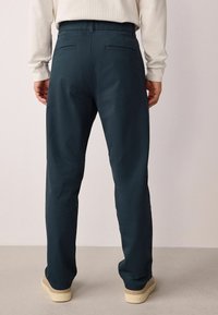 Pantalons bleu canard avec une coupe droite, dotés de deux poches arrière et d'une texture de tissu lisse. Portés avec des chaussures beiges.
