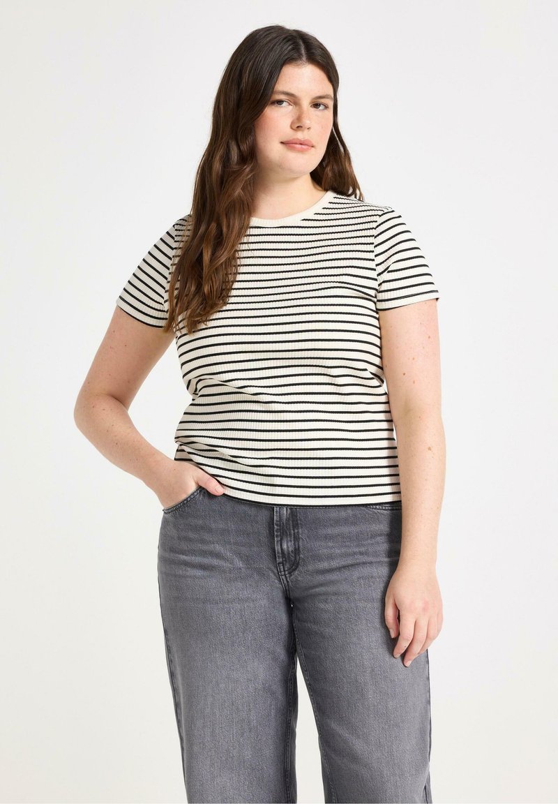 Stribet kortærmet bluse i creme og sort med ribbet tekstur. Sættes sammen med højttaljede grå jeans, med hænderne i lommerne.