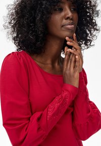 Kobieta z kręconymi włosami, ubrana w czerwoną bluzkę z długim rękawem i haftowanymi mankietami, delikatnie dotyka podbródka i palców przy twarzy.