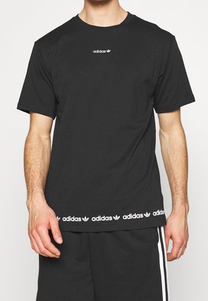 Mann trägt ein schwarzes Adidas-T-Shirt mit weißem Logo auf der Brust und wiederholten Logos am Saum, kombiniert mit schwarzen Shorts mit weißen Streifen.
