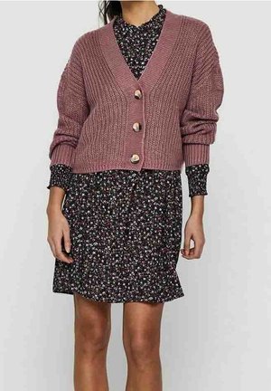 Kvinde iført en lillarosa chunky strikket cardigan med store knapper over en sort blomsterkjole med lange ærmer og høj hals.