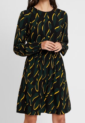 Žena oblečená v černých šatech s abstraktními zelenožlutými vzory listů, dlouhými rukávy a elastickým pasem, ruce sepnuté vpředu.